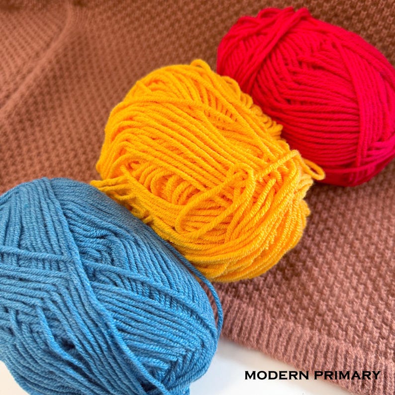 May include: Three balls of yarn in bright colors: red, yellow, and blue. The yarn is arranged on a brown knitted fabric. The text "MODERN PRIMARY" is visible in the bottom right corner.