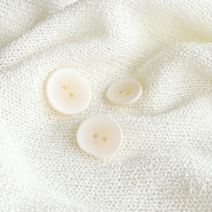 Può includere: Tre bottoni biancastri, leggermente irregolari, con due fori ciascuno, su un tessuto biancastro strutturato. I bottoni sono di dimensioni diverse, con un bottone piccolo, medio e grande.