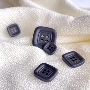 May include: Four black square buttons with four holes each. The buttons are on a white fabric background.