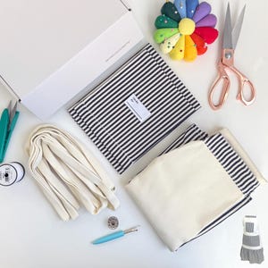 May include: A DIY kit for making a striped fabric apron. The kit includes two pieces of striped fabric, a piece of white fabric, a roll of white bias tape, a small spool of thread, a needle, a thimble, and a pincushion shaped like a flower.