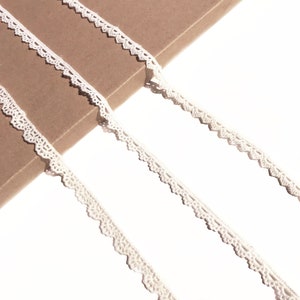 May include: White cotton lace trim with a scalloped edge. The lace is shown on a brown surface.