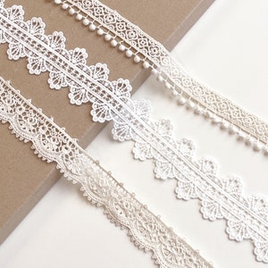 May include: Three white lace trims with different patterns. The top trim has small white pom-poms along the edge. The middle trim has a scalloped edge with a delicate floral pattern. The bottom trim has a more intricate floral pattern.