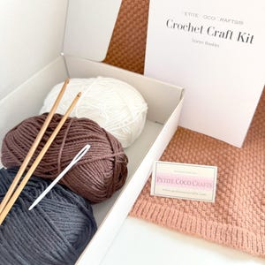 May include: A crochet craft kit in a white box, including balls of white, brown, and dark gray yarn, two wooden crochet hooks, and a white needle. The kit also includes a booklet and a small card with the text "PETITE COCO CRAFTS".