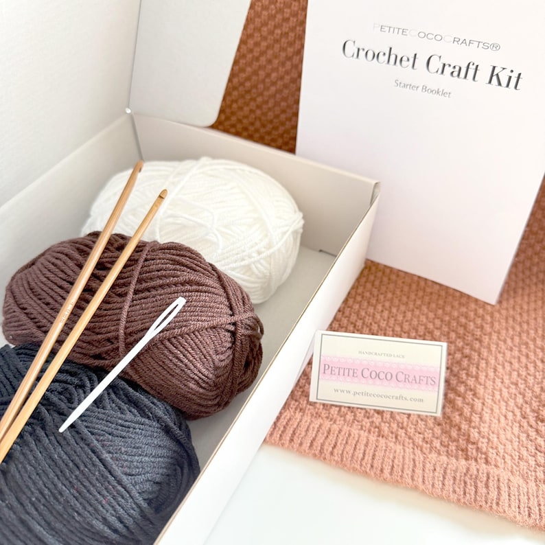 May include: A crochet craft kit in a white box, including balls of white, brown, and dark gray yarn, two wooden crochet hooks, and a white needle. The kit also includes a booklet and a small card with the text "PETITE COCO CRAFTS".