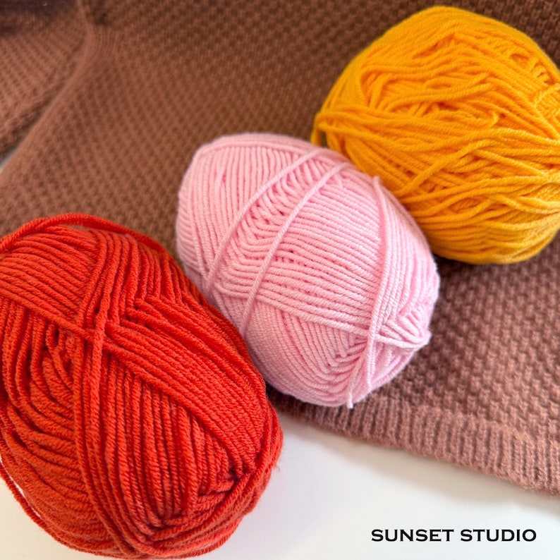 May include: Three balls of yarn in shades of red, pink, and yellow are arranged on a brown knitted surface. The yarn is tightly wound, and the colors are vibrant. The image includes the text "SUNSET STUDIO" in the bottom right corner.