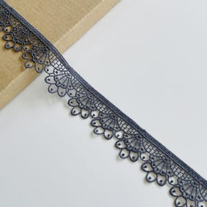 May include: Close-up of a length of dark gray lace trim. The lace features a scalloped edge with a repeating floral pattern. The trim is lying diagonally across a white surface, with a portion of a brown paper visible in the background.