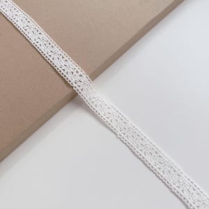 May include: Close-up of a white lace ribbon with a floral design. The ribbon is made of a delicate, open-weave fabric. The ribbon is approximately 1 inch wide and is displayed against a neutral background.