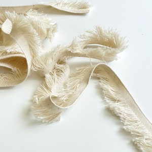 Puede incluir: Primer plano de un ribete de flecos de algodón de color crema. El fleco es suave y esponjoso, y el ribete es perfecto para añadir un toque de textura y detalle a cualquier proyecto.