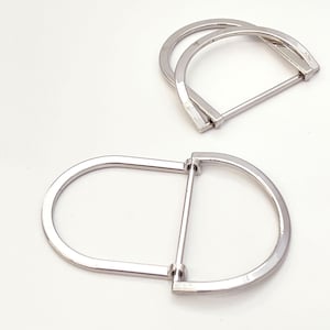 Puede incluir: Dos hebillas plateadas en forma de D. Una hebilla está abierta y la otra cerrada. Las hebillas son de metal brillante y están sobre un fondo blanco. Probablemente se utilizan para bolsos, cinturones u otros accesorios.