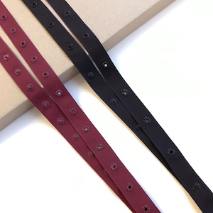 Puede incluir: Dos largos de cinta elástica con ojales metálicos. Una cinta es burdeos y la otra es negra.