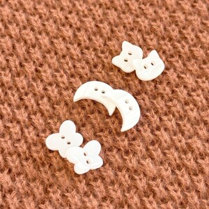 May include: A collection of white, decorative buttons in various shapes, including cat faces, crescent moons, and four-leaf clovers. The buttons are scattered on a textured, brown surface, creating a playful and whimsical aesthetic. These buttons are ideal for sewing and craft projects.