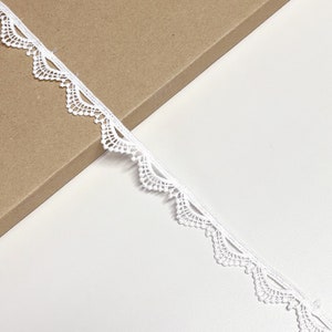May include: A length of white lace trim with a scalloped edge. The delicate lace is displayed against a white and brown background. This trim is suitable for various craft and sewing projects.