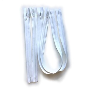 May include: Several white zippers of varying lengths are arranged against a white background. The zippers have silver metal pulls and teeth. The fabric tape is a bright white color. The zippers are for sewing and crafting projects.