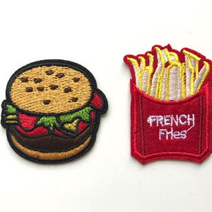 Pode incluir: Dois patches bordados para passar a ferro, um de um hambúrguer e o outro de batatas fritas. O patch do hambúrguer é castanho, vermelho e verde. O patch das batatas fritas é amarelo, vermelho e branco com o texto "French Fries".