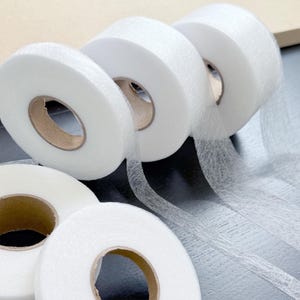 Pode incluir: Quatro rolos de fita adesiva fusível branca para artesanato de tecido. A fita está em bobinas com um centro de cartão castanho.