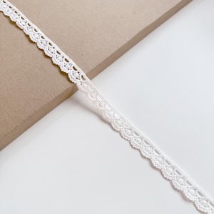 Könnte beinhalten: Ein Stück weißer Spitzenbesatz mit gewelltem Rand. Die zarte Spitze weist ein sich wiederholendes Blumenmuster auf und wird diagonal vor einem neutralen Hintergrund dargestellt. Dieser Besatz eignet sich für verschiedene Bastel- und Nähprojekte.