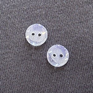 May include: Two clear, round buttons with two holes each. The buttons have a subtle iridescent sheen and are set against a dark, textured background. These buttons are suitable for sewing projects.