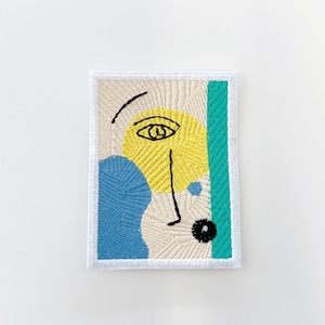 May include: Embroidered patch featuring an abstract face design. The patch has a cream-colored background with yellow, blue, and teal accents. The face is outlined in black, with a yellow circle for the eye and a blue shape for the cheek.
