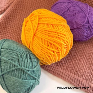 May include: Close-up of colorful yarn balls in shades of yellow, purple, and teal. The yarn is arranged on a textured, knitted fabric in a muted rose color. The text "WILDFLOWER POP" is visible at the bottom.