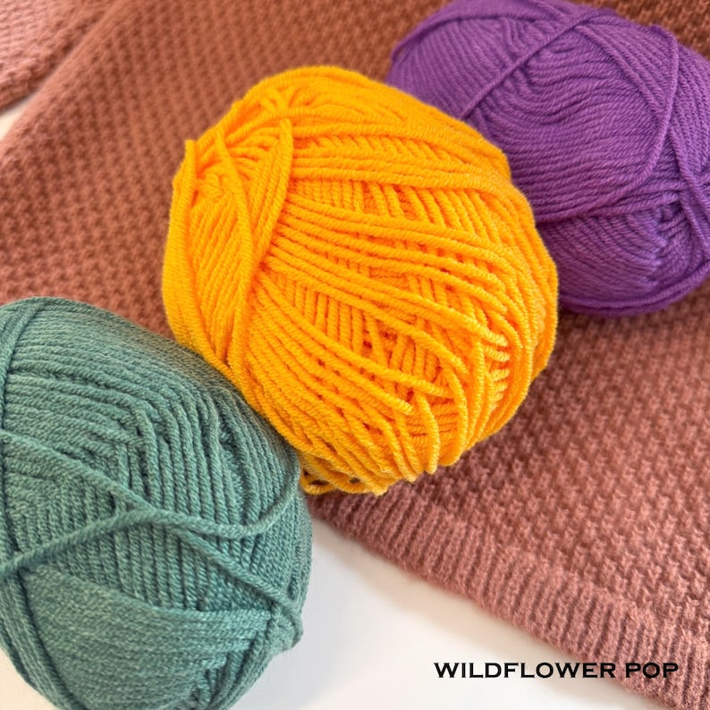 May include: Close-up of colorful yarn balls in shades of yellow, purple, and teal. The yarn is arranged on a textured, knitted fabric in a muted rose color. The text "WILDFLOWER POP" is visible at the bottom.