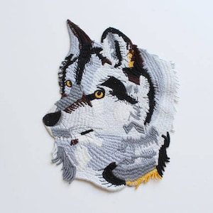 May include: An embroidered iron-on patch featuring a wolf's head with grey fur, black eyes, and a brown nose. The wolf's head is facing the left side of the image.