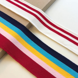 Könnte beinhalten: Nahaufnahme von bunten Gummibändern. Ein Band ist weiß mit zwei roten Streifen. Das andere Band hat Streifen in Rot, Rosa, Gelb, Blau und Marine. Diese Bänder werden wahrscheinlich für Kleidung oder Bastelarbeiten verwendet.