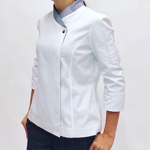 Puede incluir: Chaqueta de chef blanca con cuello mandarín y cierre de un solo botón. La chaqueta tiene un cuello de mezclilla azul contrastante y un bolsillo de parche en la manga izquierda.