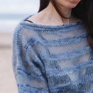Può includere: Un maglione blu chiaro a trama aperta con scollo a barchetta. Il maglione presenta strisce orizzontali in varie tonalità di blu. La maglia appare morbida e leggermente trasparente, adatta per la stratificazione. Lo sfondo è una scena di spiaggia sfocata.