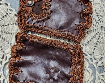 May include: A pair of brown leather fingerless gloves with intricate crochet trim. The gloves feature a button detail and a decorative crochet design on the back of the hand. The crochet is a darker shade of brown, creating a contrast with the leather.