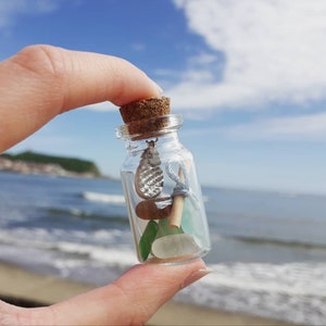 Może przedstawiać: Mała szklana butelka z korkowym korkiem wypełniona szkłem morskim, małym kawałkiem drewna unoszącego się na wodzie i srebrnym wisiorkiem. Butelka jest trzymana w dłoni na tle niebieskiego nieba i oceanu.