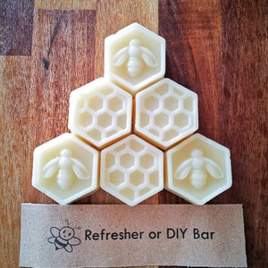May include: Six white honeycomb-shaped soap bars arranged in a pyramid shape. Each bar has a bee design on one side. The bars are on a wooden surface with a brown paper tag that reads "Refresher or DIY Bar" and has a bee illustration.