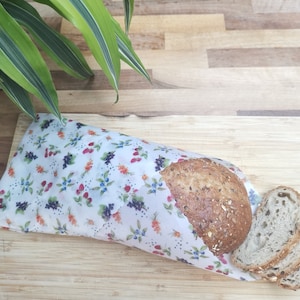 May include: A white fabric food wrap with a floral pattern featuring red berries, blue flowers, and orange leaves. The wrap is folded around a loaf of multigrain bread with a few slices cut off.