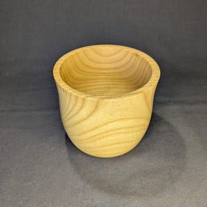 Op de afbeelding: Een kleine, ronde houten kom in een lichtbruine kleur met een opvallend, natuurlijk houtnerfpatroon. De kom heeft een gladde, gebogen buitenkant en een brede, open bovenkant, wat de organische textuur en de ringen van het hout laat zien.