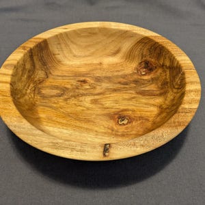 Puede incluir: Un cuenco de madera hecho a mano con un acabado natural. El cuenco es redondo con poca profundidad y presenta patrones de vetas de madera y nudos visibles. El color es marrón claro cálido.