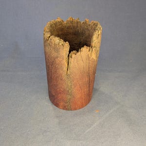 May include: A cylindrical wooden vessel featuring a natural, rough-edged rim and a deep vertical crack extending from the top. The exterior displays a rich, reddish-brown wood grain with darker, weathered textures near the jagged opening.