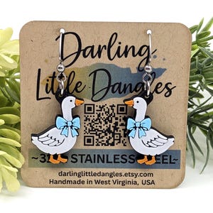 May include: A pair of goose-shaped earrings with a white body, black head, and orange beak. Each goose has a light blue bow tie. The earrings are on a brown card with the text "Darling Little Dangles" and "Stainless Steel".