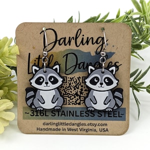 May include: A pair of raccoon earrings with a gray, white, and black design. The earrings are hanging from a brown card with the text "Darling Little Dangles" and "~316L STAINLESS STEEL~". The earrings are handmade in West Virginia, USA.