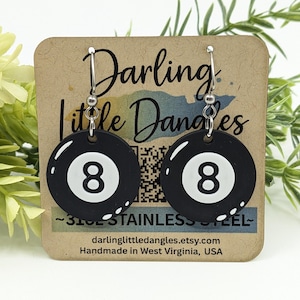 May include: A pair of black and white 8-ball earrings. The earrings are round with a black background and a white number 8. The earrings are hanging from silver hooks. The earrings are displayed on a brown card with text.