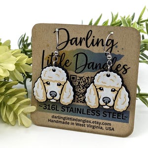 May include: Poodle earrings with a cream-colored face and fluffy fur, hanging from silver hooks. The earrings are displayed on a brown card with the text "Darling Little Dangles" and "~316L STAINLESS STEEL."