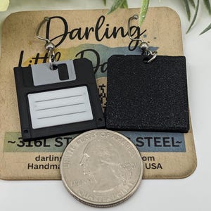 May include: Black and gray floppy disk earrings with silver-tone ear wires. The earrings are handmade and feature a retro design. The earrings are displayed on a brown card with the text "Darling Little Darlings".