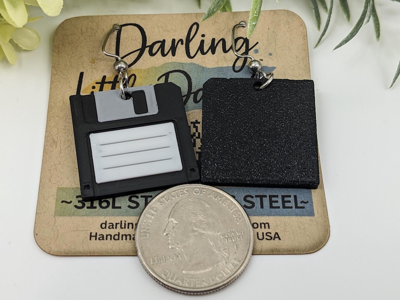 May include: Black and gray floppy disk earrings with silver-tone ear wires. The earrings are handmade and feature a retro design. The earrings are displayed on a brown card with the text "Darling Little Darlings".