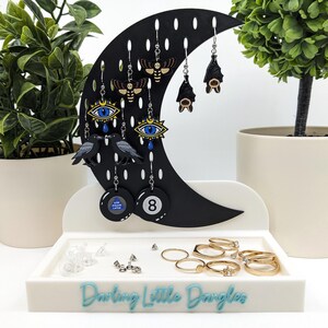 May include: A black crescent-shaped earring holder with various earrings, including bats, moths, and eyes. The holder sits on a white tray with rings and the text "Darling Little Dangles". Two potted plants flank the display.