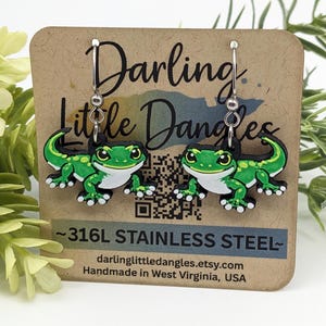 May include: A pair of gecko earrings with green and white details, hanging from silver hooks. The earrings are displayed on a brown card with the text "Darling Little Dangles" and "316L STAINLESS STEEL".