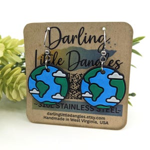 May include: Earth-shaped earrings with a blue ocean, green continents, and white clouds. The earrings are made of stainless steel and are displayed on a brown card with the text "Darling Little Dangles".