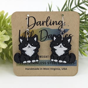 May include: A pair of dangle earrings featuring black and white cat charms with silver-toned ear wires. The earrings are displayed on a tan card printed with the text Darling Little Dangles, lightweight, stainless steel earwires, and Handmade in West Virginia, USA.