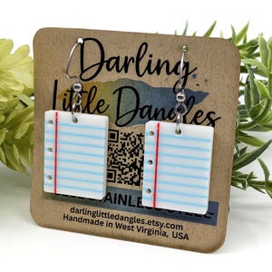 May include: White rectangular earrings with a lined paper design, featuring blue lines and a red margin. The earrings are suspended from silver-colored hooks. The earrings are on a brown card with the text "Darling Little Dangles".