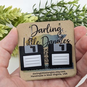 May include: A pair of black floppy disk earrings with silver-tone hooks. Each earring features a white label area and a gray slider. The earrings are displayed on a brown card with the text "Darling Little Dangles".