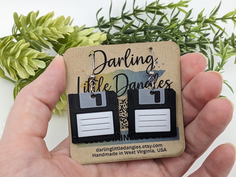 May include: A pair of black floppy disk earrings with silver-tone hooks. Each earring features a white label area and a gray slider. The earrings are displayed on a brown card with the text "Darling Little Dangles".