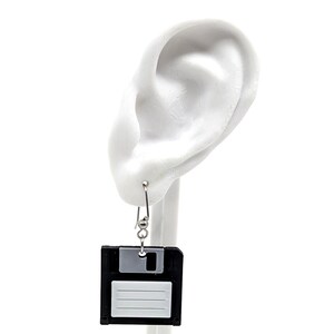 May include: A pair of novelty earrings featuring a black and gray floppy disk design. The earrings hang from silver-colored hooks, offering a unique and retro aesthetic. The earrings are a fun accessory for those who appreciate vintage tech.