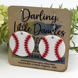 May include: White baseball earrings with red stitching, hanging from silver hooks. The earrings are displayed on a brown card with the text "Darling Little Dangles" and the Etsy shop URL. Handmade in West Virginia, USA.